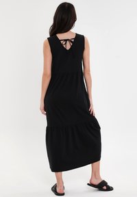 Czarna sukienka bez rękawów z warstwowym designem, zamknięciem na wiązanie z tyłu i miękkiego materiału. Długość do kostek z luźnym krojem, w połączeniu z czarnymi klapkami.