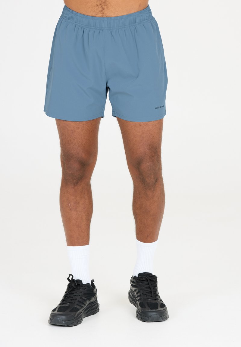 Blå sportshorts, lette, med elastisk talje. Har et diskret brandlogo på den nederste del af benet. Kombineret med sorte sneakers og hvide sokker.