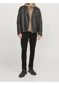 Man draagt een zwarte leren motorjack over een beige hoodie, zwarte strakke jeans en zwarte veterlaarzen, staand tegen een effen achtergrond.