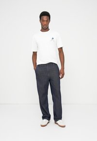 Λευκό κοντομάνικο μπλουζάκι με μπλε λογότυπο, συνδυασμένο με σκούρο τζιν παντελόνι και λευκά αθλητικά παπούτσια με καφέ λεπτομέρειες. Απλός και καθημερινός σχεδιασμός.