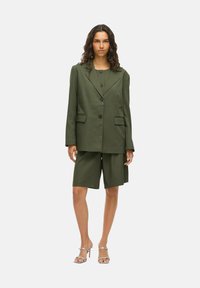 Blazer cintré vert olive avec des revers structurés, associé à un short assorti à la longueur du genou. Présente des boutons noirs et des poches épurées.
