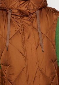 Doudoune sans manches matelassée marron avec cordons de serrage, portée par-dessus un vêtement vert à manches longues.
