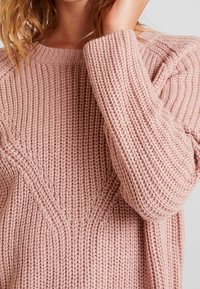 Hellrosa gestrickter Pullover mit strukturierter Musterung, geripptem Rundhalsausschnitt und überschnittenen Schulternähten. Auffallend wärmend und grob gestrickt.