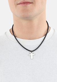 Ciondolo a forma di croce in argento su una collana di corda intrecciata nera, indossato sopra una camicia bianca con texture. La croce presenta una finitura lucida.