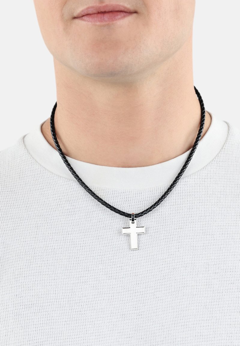 Ciondolo a forma di croce in argento su una collana di corda intrecciata nera, indossato sopra una camicia bianca con texture. La croce presenta una finitura lucida.