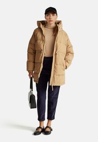 Doudoune beige avec capuche, dotée de zips avant et de poignets élastiques. Portée sur un col roulé beige, associée à un pantalon en velours côtelé bleu marine et des ballerines noires.