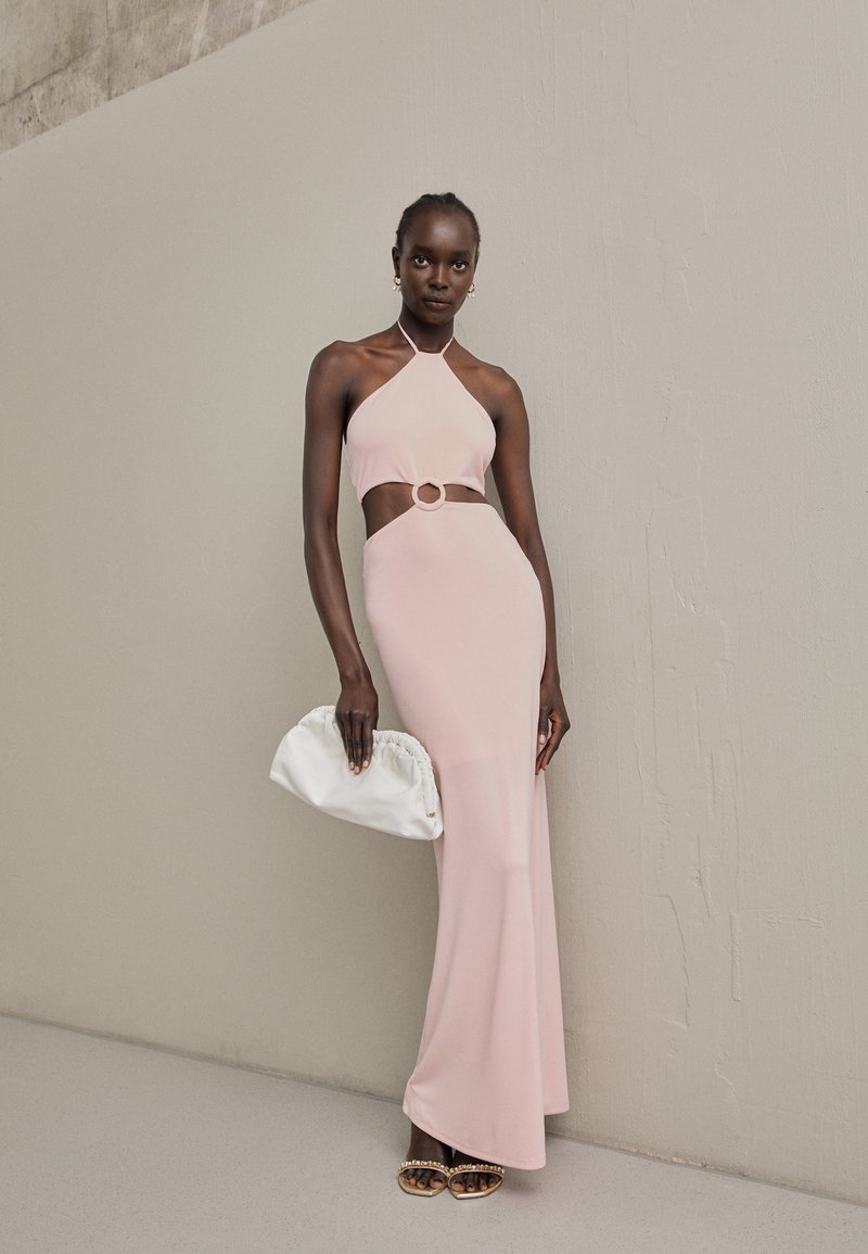 Vestido halter rosa pálido com detalhe em recorte e acento circular na cintura. Modelo segura uma clutch branca e usa sandálias adornadas.