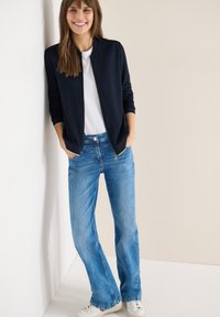 Femme souriante portant une veste zippée bleu marine, un t-shirt blanc, un jean bleu et des baskets blanches, debout de manière décontractée avec les mains dans les poches.