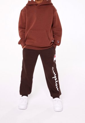 Brown hooded sweatshirt with a front pocket, paired with dark brown sweatpants featuring white "champion" text and logo, worn with white sneakers.