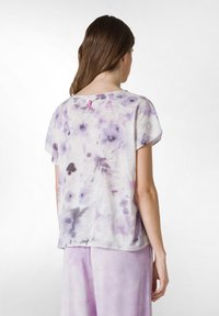 Top de tela ligera, de mangas cortas, en blanco con patrones de tie-dye morados, que presenta un escote redondo y una pequeña etiqueta rosa en la parte de atrás.