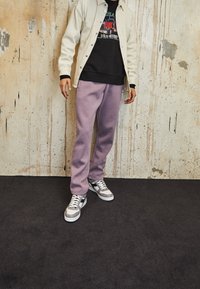 Chaqueta beige clara sobre un sudadera gráfica negra, combinada con pantalones de chándal lavanda y zapatillas grises con acentos blancos. Fondo texturizado.