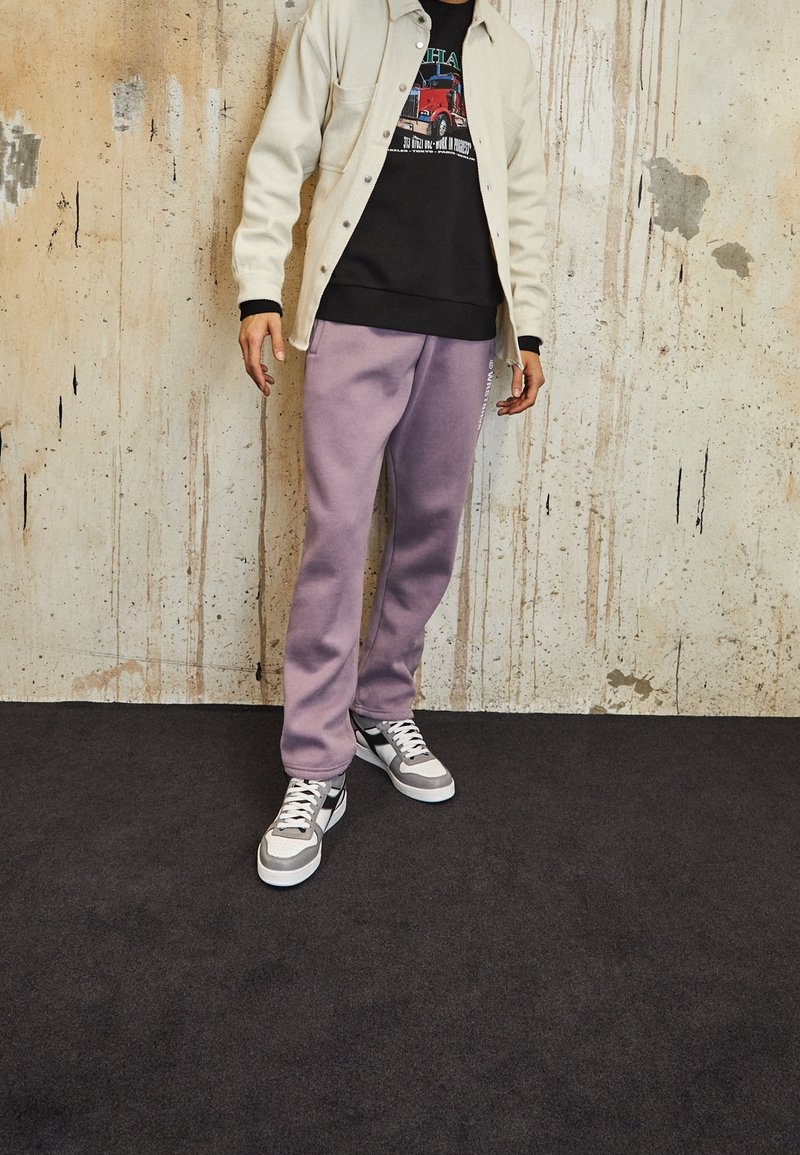 Chaqueta beige clara sobre un sudadera gráfica negra, combinada con pantalones de chándal lavanda y zapatillas grises con acentos blancos. Fondo texturizado.