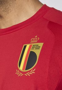 Rdeča športna majica s krožnim izrezom, z zlatim in črnim grbom, ki vključuje besedilo "ROYAL BELGIAN FA 1895" in detajl krone.