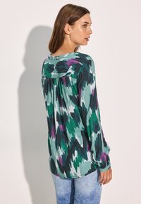 Blusa a maniche lunghe in verde, nero e viola con un motivo astratto, caratterizzata da una vestibilità morbida e un dettaglio arricciato sulla schiena. Tessuto morbido e testurizzato.