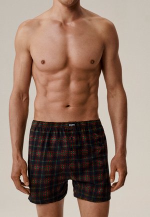 Mężczyzna bez koszulki, ubrany w ciemne, kratowane bokserki w czerwone i zielone paski, stojący na jednolitym tle.