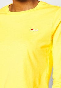 Gul bomullströja med långa ärmar och rundad halsringning, med en liten broderad röd och vit logotyp på vänster bröst. Slät textur.