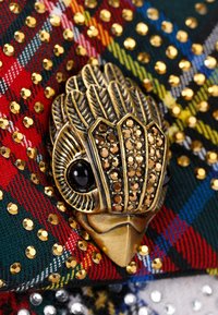 Spilla di bronzo a forma di uccello con dettagli intricati delle piume e accenti in cristallo, posizionata su un tessuto tartan multicolore arricchito da paillettes.