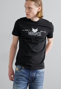 Juodos medvilnės marškinėliai su baltu Versace logotipu ir tekstu, apvaliu kaklu, trumpomis rankovėmis ir prigludusia forma, derinami su šviesiai mėlynomis džinsomis.