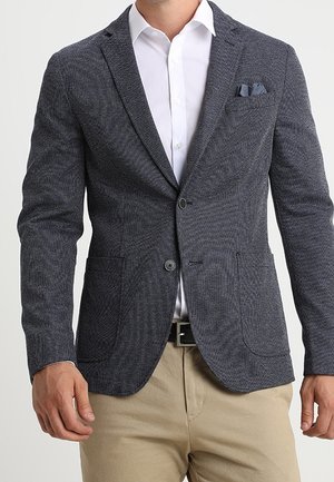 Mann trägt ein tailliertes, dunkelgraues, strukturiertes Sakko über einem weißen Hemd mit Knöpfen und beigen Hosen, mit einem grauen Einstecktuch im Sakko.