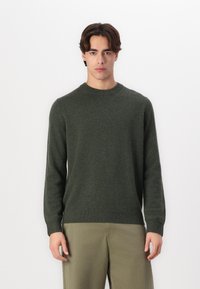Mørkegrønn strikkegenser med rund hals, ribbede mansjetter og hem, med en myk tekstur. Kombinert med lyse khaki bukser.