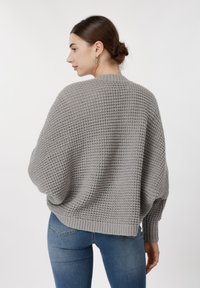 Γυναίκα με τα μαλλιά της σε κότσο, φορά ασημένια σκουλαρίκια κρίκους, ένα χαλαρό γκρι πλεκτό πουλόβερ και μπλε τζιν, σε πλαϊνή όψη απέναντι σε λευκό φόντο.