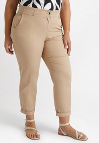 Beige, avsmalnande byxor med uppvikt nederkant, tillverkade av slät tyg. Har framfickor och knäppning med knappar. Bärs med sandaler med remmar.