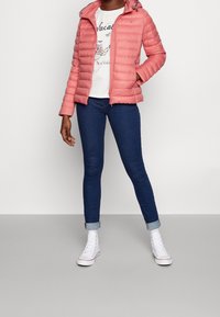 Doudoune rose avec couture horizontale, fermeture éclair, capuche à cordon de serrage. Associée à un t-shirt blanc à motif, un jean skinny bleu foncé et des baskets blanches.