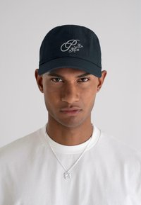 Navy blue baseball cap with embroidered logo and floral design. The model wears a white crewneck shirt and silver chain necklace.