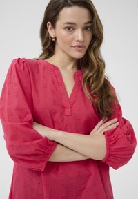 Blouse fuchsia avec un décolleté en V, broderie en forme de cœur et manches bouffantes élastiquées. Tissu léger et coupe décontractée.