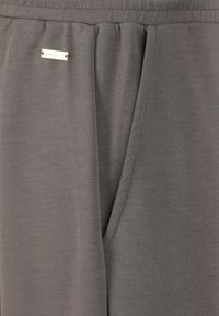 Graue Sporthose aus weichem Stoff mit einem breiten Bund, ausgestattet mit einer Seitentasche und einem kleinen metallischen Logoanhänger.