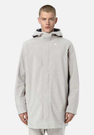 Giovane uomo che indossa un cappotto con cappuccio grigio chiaro con un piccolo logo sul petto, in piedi davanti a uno sfondo bianco semplice.