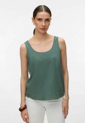 Ženska z rjavimi lasmi, zvezanimi nazaj, oblečena v zeleno brezrokavnik in bele hlače, stoji pred belim ozadjem.