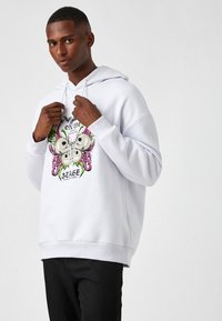 Sudadera blanca con un colorido estampado gráfico de calaveras y acentos florales en la parte delantera. La tela parece suave y tiene un corte relajado.