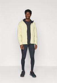 Světle žlutá kapucňová bunda, černé tričko, černé legíny a černé sportovní boty. Hladká tkanina a minimalistický design.