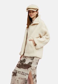 Chaqueta de forro polar color crema con cierre frontal, ajuste oversize y bolsillos laterales, combinada con una falda estampada que presenta gráficos intrincados.