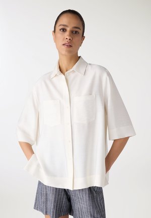 Donna che indossa una camicia bianca a maniche corte, ampia, con due tasche sul petto e pantaloncini grigi a righe sottili, in piedi con le mani nelle tasche.