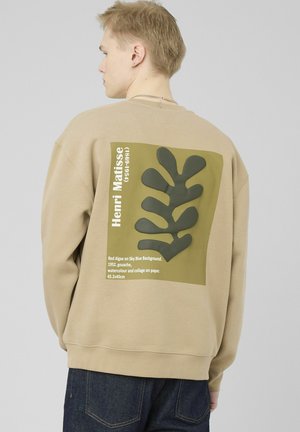 Jeune personne portant un sweat-shirt beige avec une illustration abstraite de feuille verte et le texte Henri Matisse au dos, debout devant un fond gris clair.