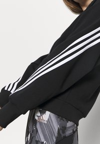 Sudadera negra con diseño de tres rayas blancas en las mangas, con cierre de cremallera en la parte delantera, puños acanalados y una textura suave, llevada sobre leggings estampados.