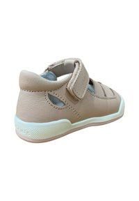 Zapato de cuero color beige claro con una cinta de velcro, recortes laterales, suela texturizada y detalles blancos. Forma redondeada con costuras decorativas.