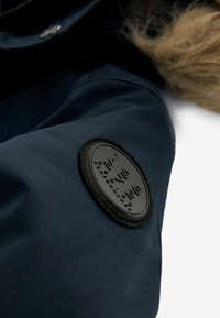 Manica di giacca impermeabile blu navy con un patch circolare nero caratterizzato da testo in rilievo e un bordo testurizzato, insieme a un bordo in pelliccia sintetica.