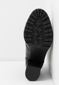 Bota de cuero negro con una suela de goma texturizada, que presenta un patrón de relieve resistente y un pequeño tacón cuadrado. El diseño es elegante y aerodinámico.