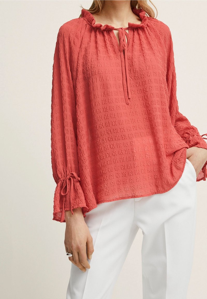Femme portant un chemisier corail texturé à manches longues avec col à volants et détails noués, associé à un pantalon blanc, main dans la poche.