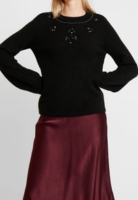 Pull en laine noir à encolure ronde, orné de perles noires et de sequins, associé à une jupe fluide en satin bordeaux.