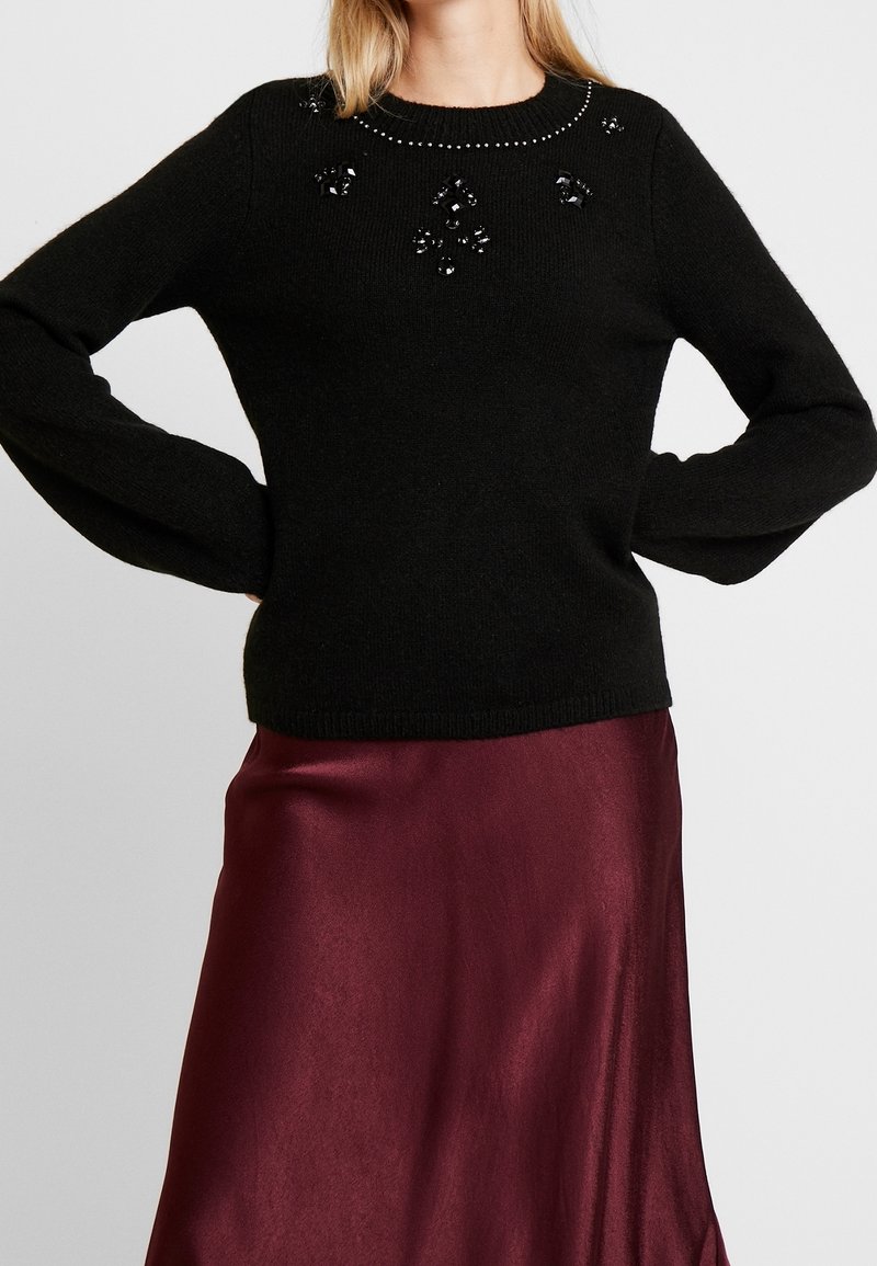 Pull en laine noir à encolure ronde, orné de perles noires et de sequins, associé à une jupe fluide en satin bordeaux.