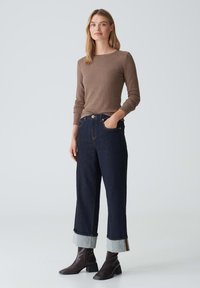 Braun gerippter langärmeliger Oberteil kombiniert mit dunkelblauen weit geschnittenen Jeans mit hochgekrempelten Säumen in Hellblau, getragen mit braunen Absatzstiefeln.