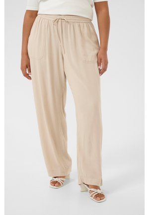 Femme portant un pantalon beige à jambes larges avec taille à cordon et poches avant, assorti à des sandales blanches à talons et à brides.