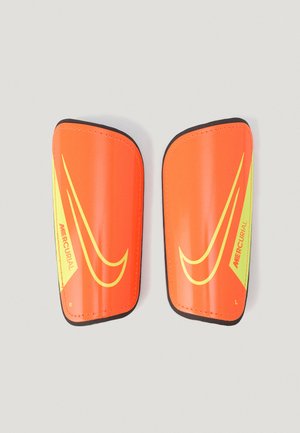 Caneleiras Nike Mercurial em laranja e amarelo, leves, com acabamento em plástico suave, bordas arredondadas e etiquetas indicando o lado esquerdo e o lado direito.