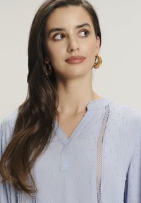 Blusa azul claro con tejido texturizado, escote en forma de V, adornos de encaje y un patrón de lunares. Mangas largas con un ajuste relajado. Pendientes de aro dorados.