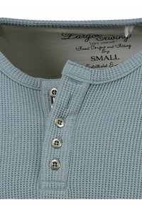 Maglietta henley azzurro chiaro con un tessuto a nido d'ape testurizzato, tre bottoni di colore argento e colletto a costine. Tag di taglia: piccola.