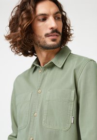 Long-sleeve green shirt made of cotton with a collar, two chest pockets, beige buttons, and a small logo tag on the side.